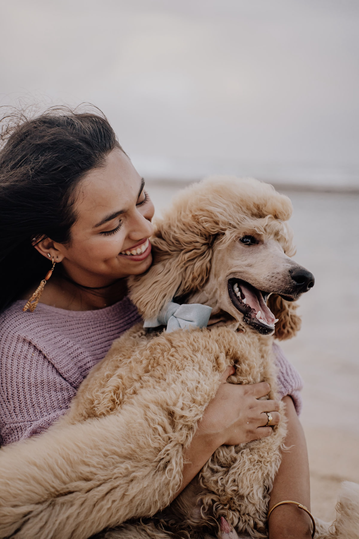Adaia sonríe y abraza a un caniche color arena con un lazo azul, en la playa